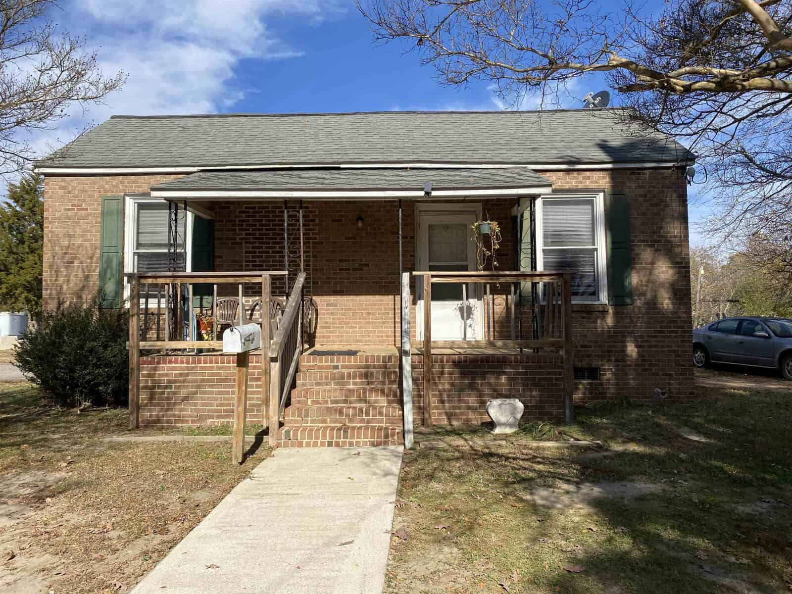 A single-story brick house with a small front porch and steps leading up to the main entrance, on a sunny day with a clear sky and a car parked in the driveway to the left.
