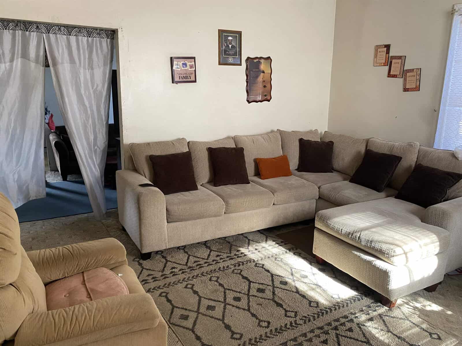 A living room with a large l-shaped sofa adorned with cushions, a matching ottoman, and a patterned area rug. the walls are decorated with various picture frames, and there's a view into another room through a curtained doorway to the left.