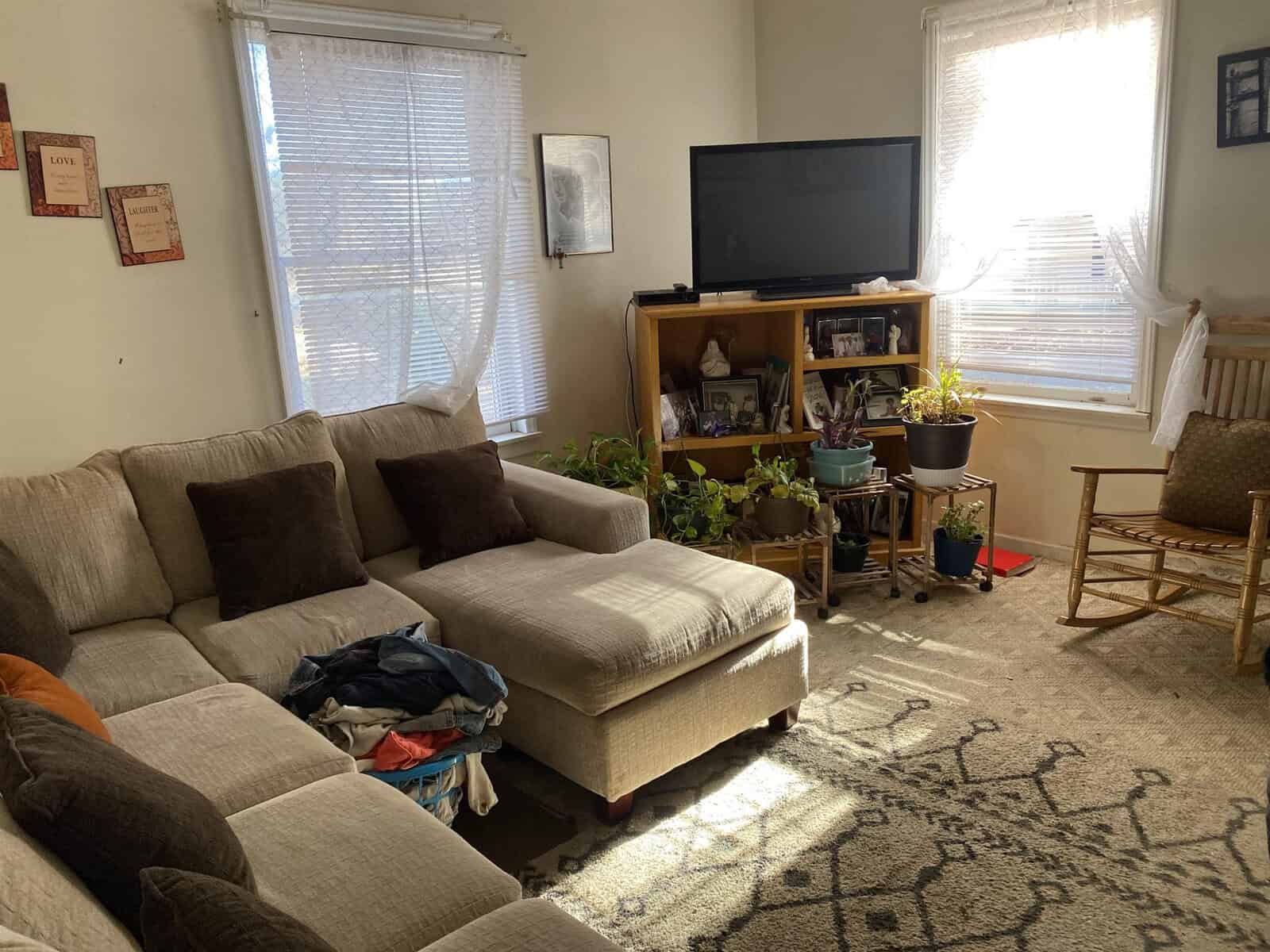 A sunlit living room with a sectional sofa and a pile of clothes on the ottoman, featuring a television stand with various decorative items, houseplants by the window, and a rocking chair to the side.