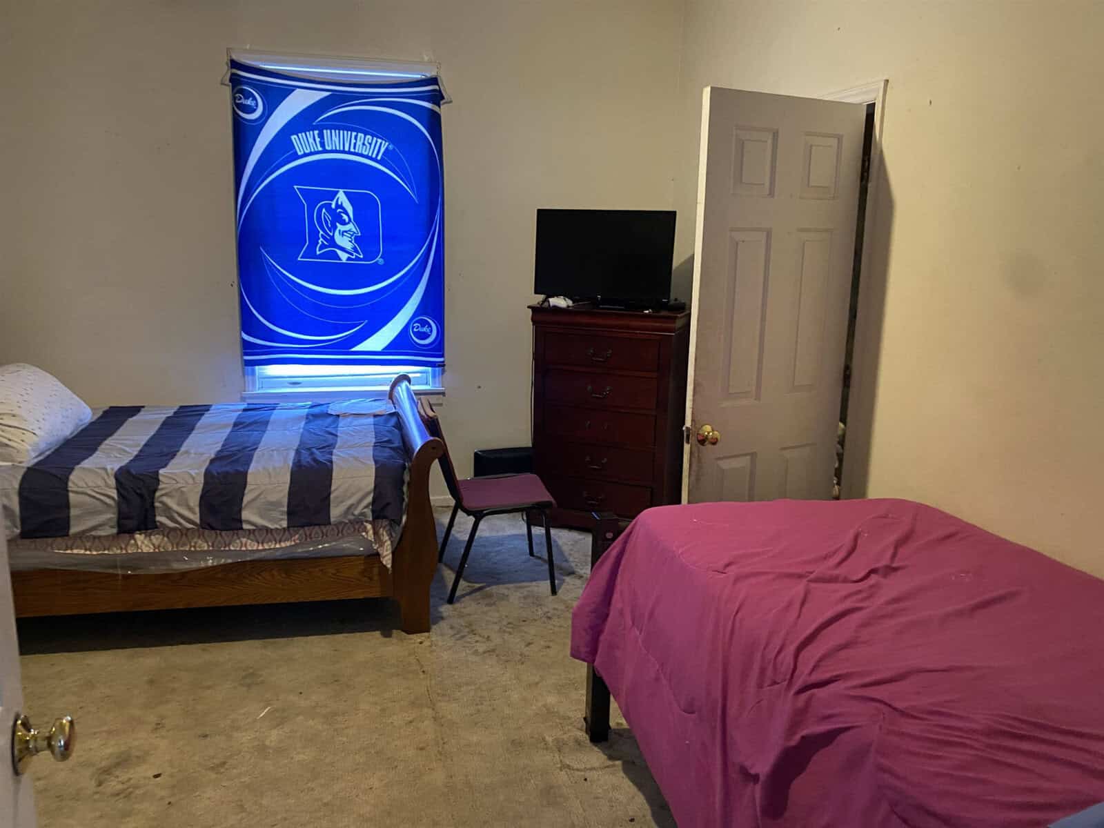 A modestly furnished room with two made beds featuring striped and solid pink bedding, a wooden dresser, a desk with a chair, a television, and a prominent duke university flag on the wall.