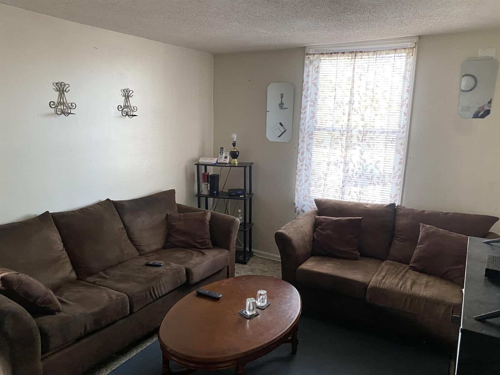 A modest living room with a brown sofa and loveseat, a wooden coffee table, sparse decorations, and natural light streaming through a curtained window.
