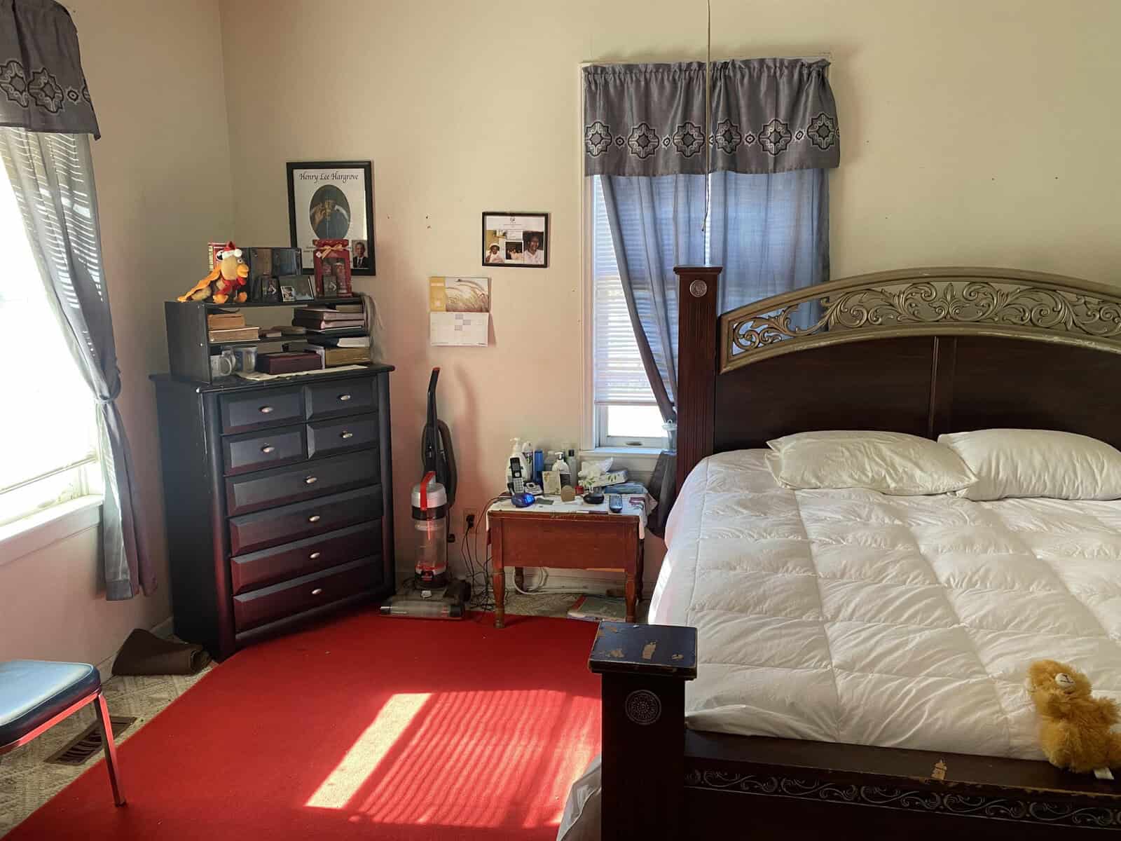 A moderately lit bedroom featuring a made bed with a white comforter, a red carpet, a dresser with a television and various items on top, a vacuum cleaner beside it, and a small blue bedside table with a lamp.