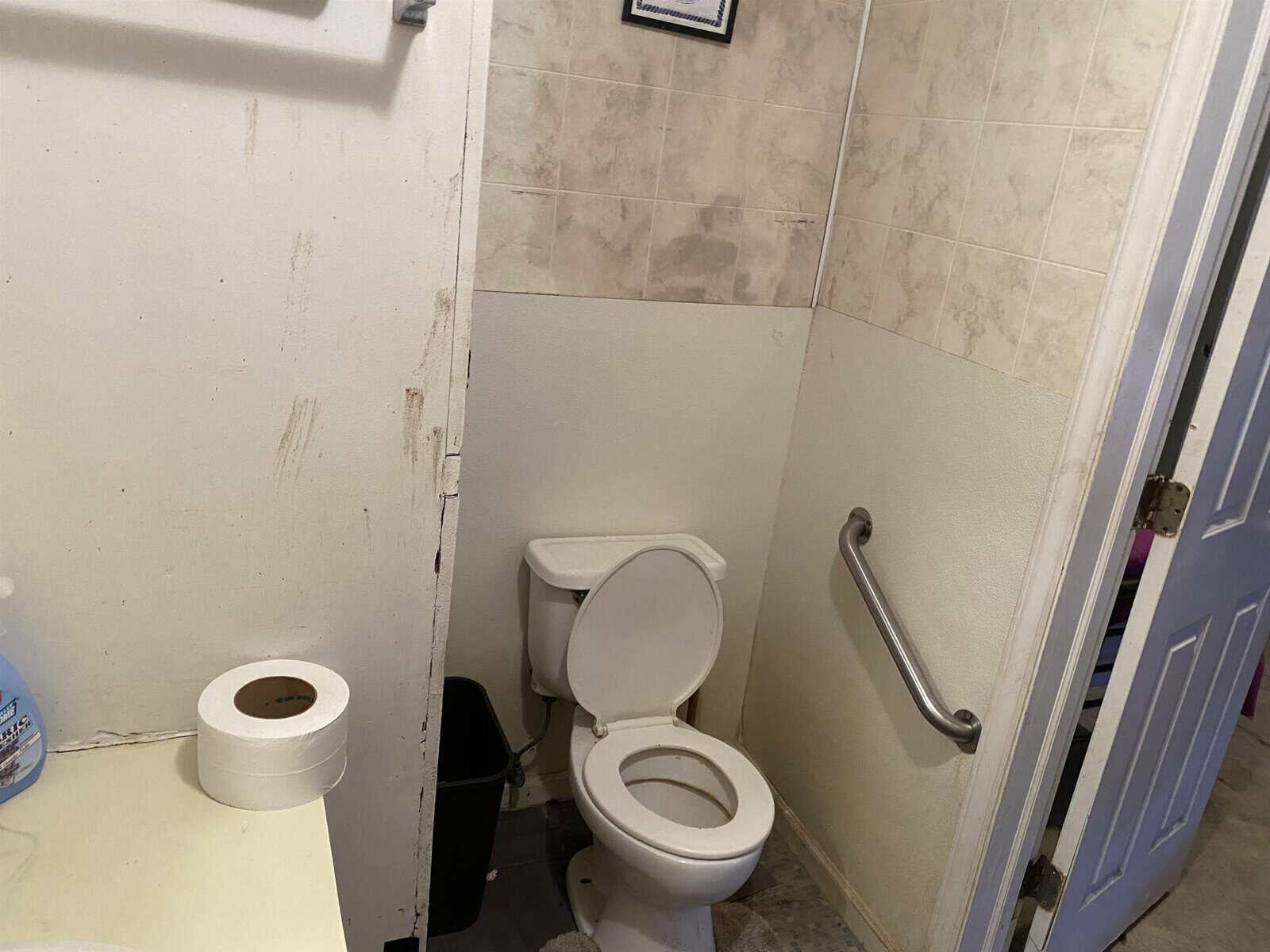 A small bathroom with white walls showing signs of wear or damage, a toilet with an open lid, a roll of toilet paper on the floor, and a grab bar installed on the wall.